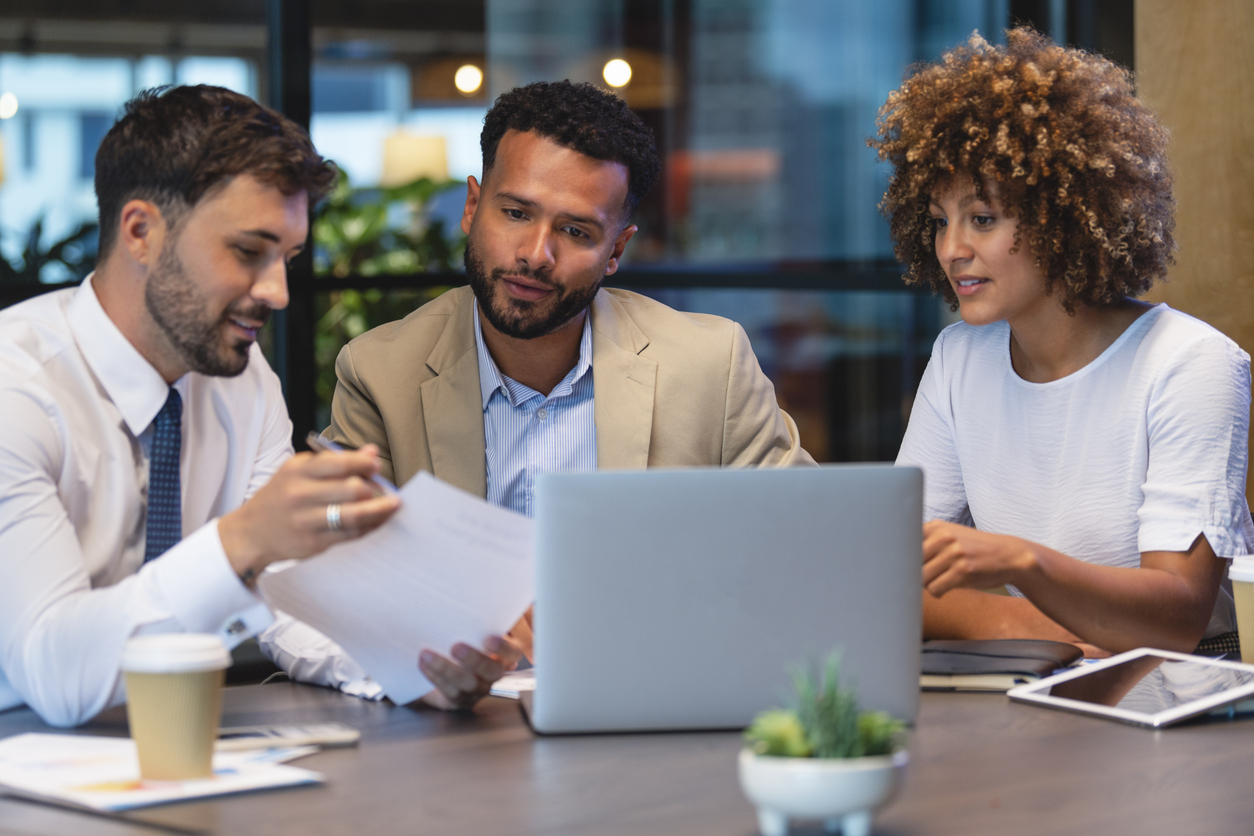 Three business people meeting and looking at a laptop and a document. There is paperwork and other technology on the table, formal business wear.